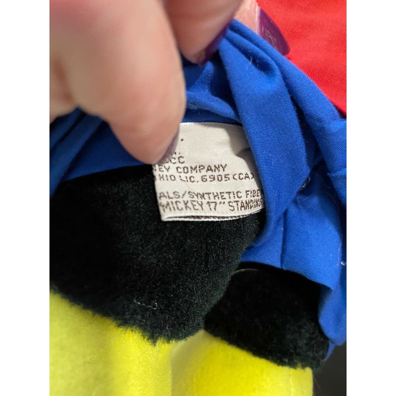 Pode incluir: Close-up da etiqueta de um brinquedo de pel&uacute;cia do Mickey Mouse. A etiqueta &eacute; branca com texto preto que diz "Disney Company, KIO LIC. 6905 (CA), ALS/SYNTHETIC FIBER, MICKEY 17" STANDING".