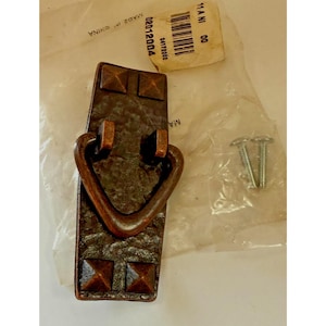 以下が含まれることがあります： テクスチャ加工された表面と正方形のリベットのディテールが特徴の、ブロンズトーンの金属製キャビネットプル。プルには湾曲したハンドルがあり、取り付けネジが含まれています。「11 A NI 00」というテキストとバーコードが付いたタグが表示されます。