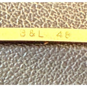 May include: A gold-toned metal bar featuring the embossed text B & L 48, positioned against a dark brown textured surface. The metallic strip displays a smooth, reflective finish with clear, legible characters centered horizontally on the surface.