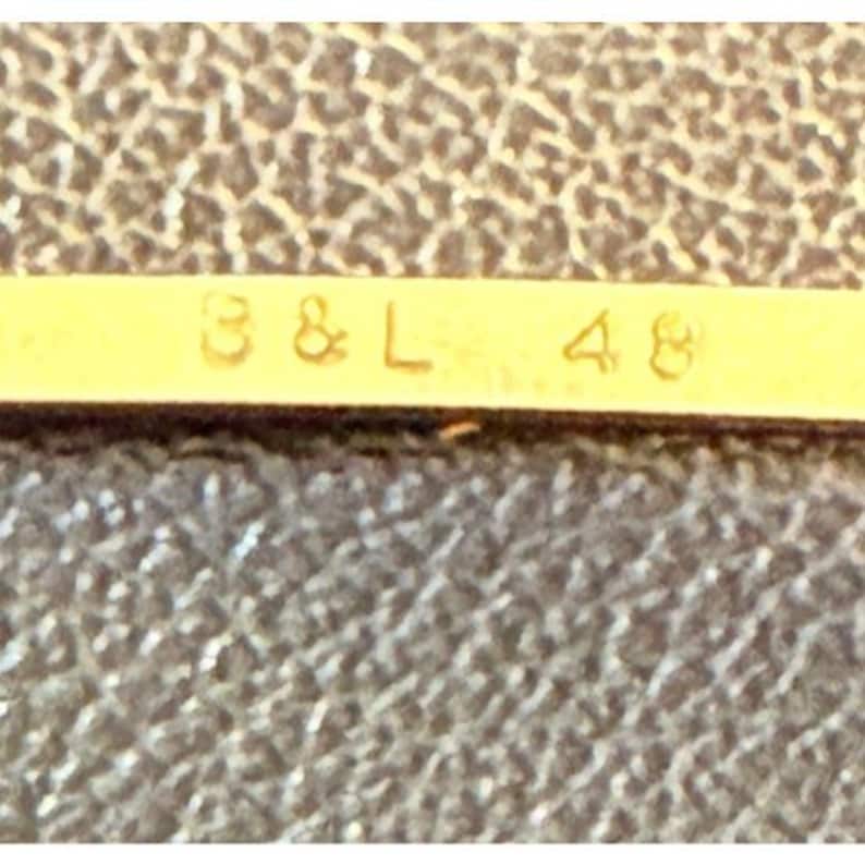 May include: A gold-toned metal bar featuring the embossed text B & L 48, positioned against a dark brown textured surface. The metallic strip displays a smooth, reflective finish with clear, legible characters centered horizontally on the surface.