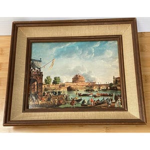 May include: A framed painting depicting a scene of a river with boats and people. The painting is set in a city with a large building in the background. The painting is framed in a brown wooden frame with a beige mat.