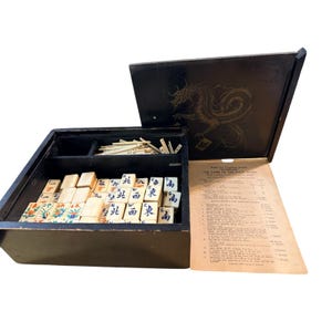 Puede incluir: Un juego de Mahjong vintage de madera negra con una ilustración de dragón en la tapa. El juego incluye fichas de color marfil con caracteres chinos y diseños florales coloridos. Se ve una hoja de reglas.
