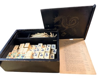 May include: A vintage black wooden Mahjong set with a dragon illustration on the lid. The set includes ivory-colored tiles with Chinese characters and colorful floral designs. A rule sheet is visible.