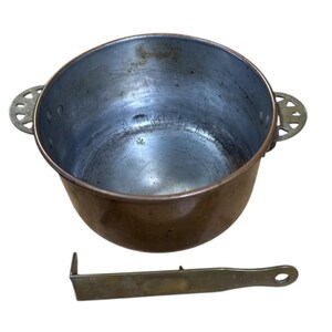 May include: A copper cooking pot with decorative handles and a metal stirring tool. The pot has a wide opening and a rounded base. The stirring tool is rectangular with a rounded handle. The pot is likely used for cooking.