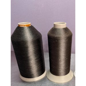 May include: Two large spools of dark charcoal gray sewing thread. Each spool features a tapered plastic core with a light gray base and top. The left spool has a brown label at the top with the white text 20000m printed clearly on the surface.