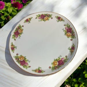 Pode incluir: Um prato de cerâmica branco com uma borda dourada e um design floral com rosas vermelhas e amarelas. O prato é perfeito para servir sobremesas ou lanches.