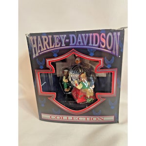 Peut inclure: Une décoration de Noël Harley-Davidson représentant deux personnages en vert et rouge jouant des instruments de musique. La décoration est dans une boîte noire avec le logo Harley-Davidson et les mots "Harley-Davidson Collection".