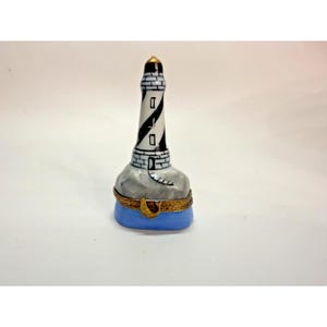 May include: A porcelain trinket box shaped like a lighthouse with a black and white spiral pattern, a grey rocky base, and a light blue bottom section. The piece features a gold-toned metal hinge and a small gold-toned metal clasp on the front.