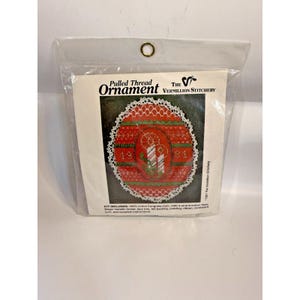 May include: A packaged craft kit for a pulled thread ornament. The ornament features a red design with a white lace trim, green accents, and the year 1981. The package is clear plastic with the text "Pulled Thread Ornament" and "The Vermillion Stitchery".