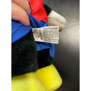Pode incluir: Um brinquedo de pel&uacute;cia preto e branco com roupas vermelhas e azuis. O brinquedo tem sapatos amarelos e uma etiqueta que diz "applause Woodland Hill Made in China The Walt Disney Co. Reg. No. PA 2910 Contents: All New Materials Item No. 14763".