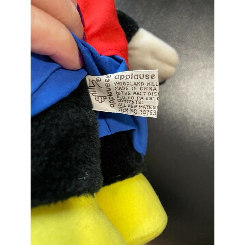Pode incluir: Um brinquedo de pel&uacute;cia preto e branco com roupas vermelhas e azuis. O brinquedo tem sapatos amarelos e uma etiqueta que diz "applause Woodland Hill Made in China The Walt Disney Co. Reg. No. PA 2910 Contents: All New Materials Item No. 14763".