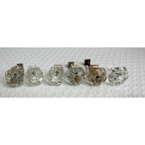 May include: A set of six clear glass cabinet knobs, each featuring a hexagonal shape with faceted edges. The knobs are arranged in a horizontal row, showing the transparent glass construction and the attached metal hardware components.