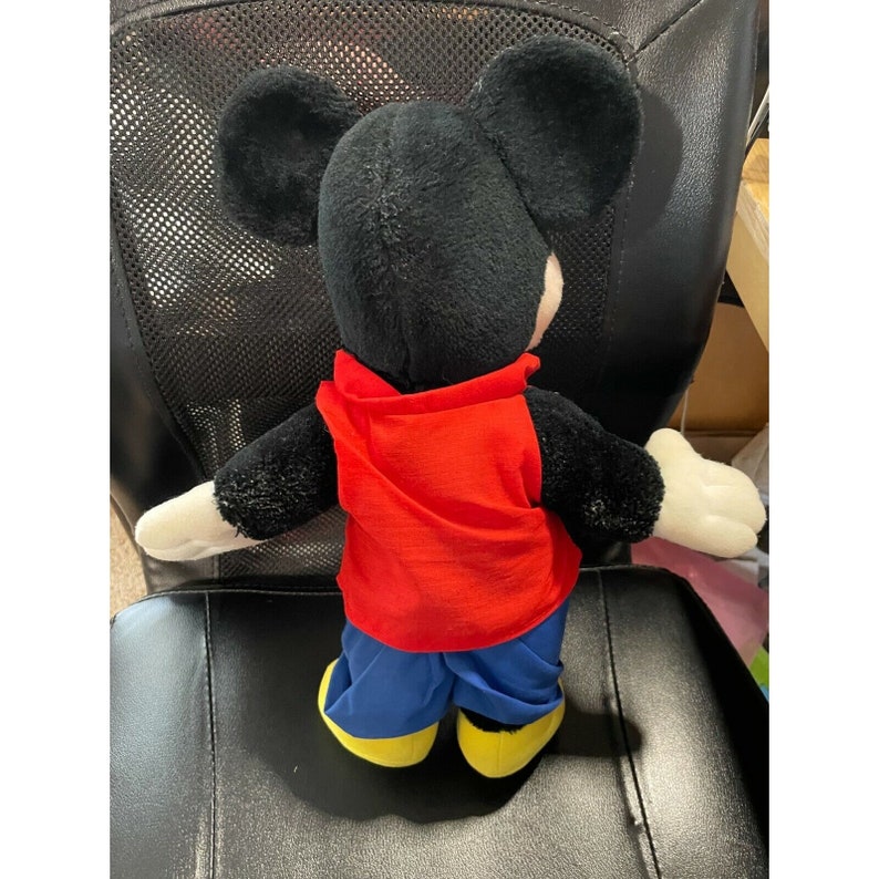 Pode incluir: Um brinquedo de pel&uacute;cia Mickey Mouse preto vestindo uma camisa vermelha e cal&ccedil;as azuis. O brinquedo tem orelhas grandes e uma express&atilde;o amig&aacute;vel.