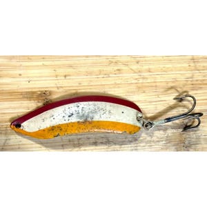 Puede incluir: Un señuelo de pesca vintage con un cuerpo metálico curvado. El señuelo presenta una combinación de colores rojo, blanco y amarillo, con un anzuelo doble adjunto. El señuelo está diseñado para atraer peces.