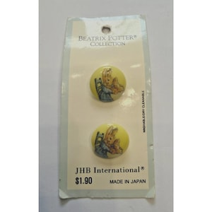 Op de afbeelding: Twee ronde gele knopen uit de Beatrix Potter Collectie van JHB International. Elke knoop heeft een gedrukte illustratie van een konijn dat op een stoel zit. De knopen zijn bevestigd op een rechthoekige kaart met de tekst Made in Japan.