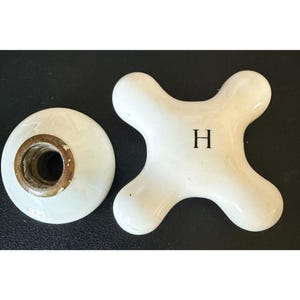 May include: A white ceramic cross-shaped faucet handle featuring the letter H in black, accompanied by a separate circular white ceramic base component with a metallic threaded interior fitting for installation on a plumbing fixture.