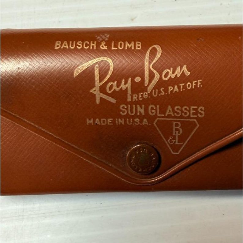 May include: A brown textured leather sunglasses case with a snap button closure. The front features gold embossed text reading BAUSCH & LOMB, Ray-Ban, REG. U.S. PAT. OFF., SUN GLASSES, MADE IN U.S.A., and a B&L logo inside a triangle shape.