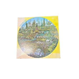 Puede incluir: Un rompecabezas circular que presenta una escena detallada de un paisaje con árboles, un arroyo y varias plantas. El rompecabezas está sobre un fondo amarillo. El texto "A Round Jigsaw Puzzle" es visible en la parte superior izquierda.