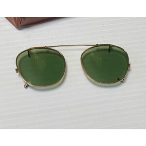 May include: A pair of clip-on sunglasses featuring round green lenses encased in thin gold-tone metal frames. The lenses are connected by a single horizontal bridge bar, with small metal clips attached to the outer edges for securing to eyewear.
