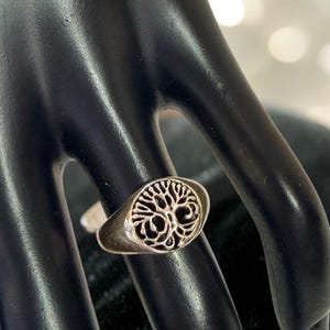 Könnte beinhalten: Ein silberfarbener Ring mit ovaler Oberfläche, der ein detailliertes Lebensbaum-Design zeigt. Der Ring ist vor einem dunklen Hintergrund platziert, der die komplizierte filigrane Arbeit des Baummotivs hervorhebt. Der Ring hat ein glattes, schlichtes Band.