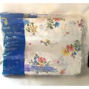 Könnte beinhalten: Ein weißes, flaches Laken mit einem Blumenmuster in Rosa-, Gelb- und Blautönen. Das Laken ist in Plastik verpackt und hat ein blaues Etikett mit der Aufschrift "Danville No Iron Sheet" und der Größe "One Double Flat Sheet for 54x75 in (137x191 cm) Mattress, Finished Size: 81x96 in (206x244 cm)".