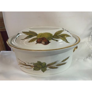 May include: White ceramic casserole dish with a lid. The dish has a gold rim and a floral design featuring green leaves, brown nuts, and wheat stalks.