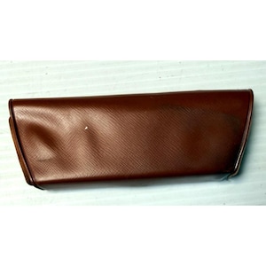 May include: A rectangular, brown, hard-shell glasses case with a textured, cross-hatched surface pattern. The case features a hinged lid design with a smooth, slightly curved front profile and tapered sides, providing a secure enclosure.