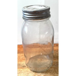 Op de afbeelding: Een glazen pot met een zilveren deksel. Het deksel heeft een verhoogde rand en een klein gaatje in het midden.