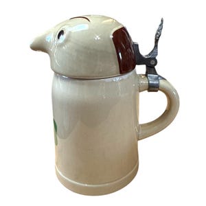 Peut inclure: Une chope à bière en céramique beige avec un couvercle en forme de chien et une fermeture à charnière en métal. La tête du chien est marron et beige, avec un motif de feuille verte sur le côté.