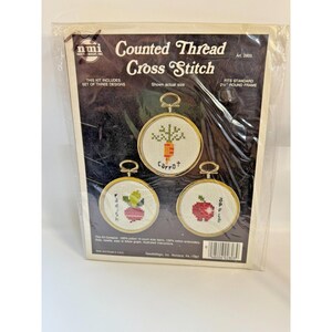 May include: A cross-stitch kit featuring three designs: a carrot, a radish, and an apple. Each design is displayed in a gold-colored round frame. The kit includes 100% cotton fabric and embroidery floss. The packaging reads "Counted Thread Cross Stitch."