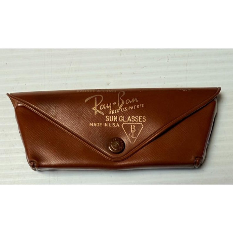May include: A brown, textured, triangular-shaped sunglasses case with a snap button closure. The front features gold-colored text reading Ray-Ban, REG. U.S. PAT. OFF., SUN GLASSES, MADE IN U.S.A., and a B&L logo inside a triangle.