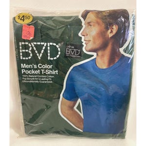 Könnte beinhalten: Eine grüne Verpackung mit einem blauen T-Shirt mit Tasche darin. Die Verpackung trägt den Text "BVD Men's Color Pocket T-Shirt 100% Natural Combed Cotton. Pre-Shrunk for a Lasting Fit. Unconditionally Guaranteed." und "L42-44 BVD 100% Combed Cotton".