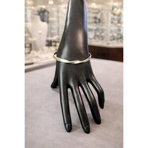 May include: A silver-colored, twisted bangle bracelet displayed on a black hand-shaped jewelry holder. The bracelet has a simple, elegant design. The background is blurred, showing a jewelry store display.