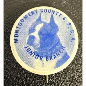 May include: A round pinback button featuring a blue and white illustration of a dog's head. The text "MONTGOMERY COUNTY S.P.C.A." is printed in blue along the top edge, and the words "JUNIOR BRANCH" are printed in blue along the bottom edge.