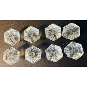 May include: Eight clear glass buttons, each shaped as a hexagon. The buttons feature a central hole and a series of white, opaque, parallel stripes radiating from the center toward the edges of the hexagonal form, creating a geometric pattern.