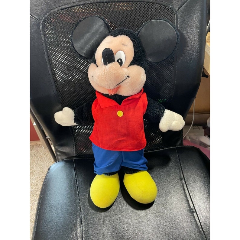 Pode incluir: Um brinquedo de pel&uacute;cia do Mickey Mouse vestindo uma camisa vermelha com um bot&atilde;o amarelo e cal&ccedil;as azuis. O brinquedo tem orelhas pretas e um nariz preto. O brinquedo est&aacute; sentado em uma cadeira preta.