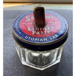 Könnte beinhalten: Ein vintage Sanford's Library Paste Utopian Jar mit einem klaren, facettierten Glasboden und einem runden Metalldeckel. Der Deckel zeigt ein rotes und blaues Design mit weißem Text, der Sanford's Library Paste liest, sowie einen zentralen Griff.