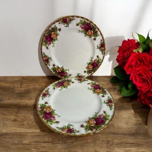 以下が含まれることがあります： 金色の縁取りと赤、ピンク、黄色のバラの花柄が施された白いセラミック製のプレートが2枚。プレートは重ねられています。