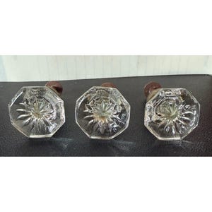 May include: Three clear glass cabinet knobs with an octagonal shape and a starburst pattern radiating from the center. Each knob features a dark, weathered metal base attachment, providing a contrast to the transparent, faceted glass surface.
