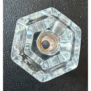 May include: A clear, hexagonal glass knob featuring a faceted, geometric design with a central circular opening. The interior of the center hole displays a metallic, copper-toned threaded insert, providing a contrast to the transparent glass body.