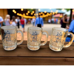 Peut inclure: Trois chopes à bière en verre transparent avec des anses sont présentées sur une surface en bois. Chaque chope présente un motif bleu et blanc d'une chope à bière traditionnelle. L'arrière-plan est flou, suggérant un cadre extérieur.