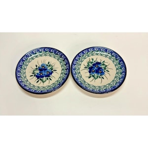 Pode incluir: Dois pires redondos de cerâmica com um centro de cor creme e um motivo floral de amores-perfeitos azuis pintado à mão. As bordas são decoradas com um padrão repetitivo de vinhas azuis e verdes sobre um fundo creme, formando um conjunto.