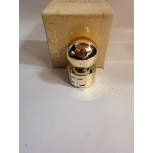 May include: A gold-colored perfume bottle with a spherical top and a cylindrical base. The base has the letters "EC" engraved on it. The bottle is in front of a beige box with the word "emprunte" printed on it.
