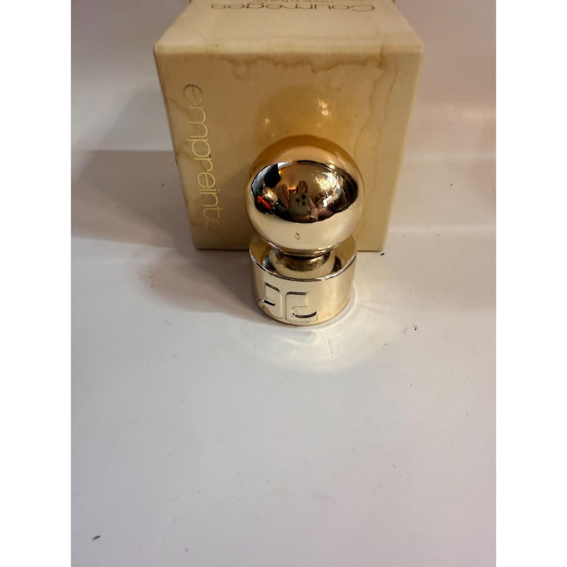 May include: A gold-colored perfume bottle with a spherical top and a cylindrical base. The base has the letters "EC" engraved on it. The bottle is in front of a beige box with the word "emprunte" printed on it.