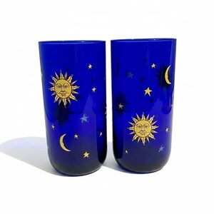 May include: Two cobalt blue drinking glasses with celestial designs. Each glass features a smiling sun, crescent moons, and stars in yellow. The glasses are cylindrical and have a gradient effect from dark blue to a lighter shade.