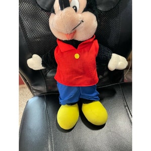 Pode incluir: Um brinquedo de pel&uacute;cia do Mickey Mouse vestindo uma camisa vermelha com um bot&atilde;o amarelo, cal&ccedil;as azuis e sapatos amarelos.