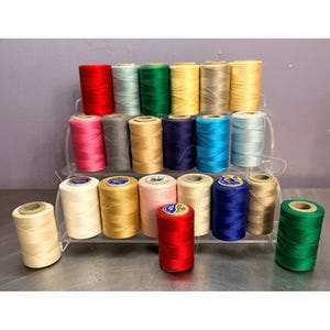 May include: A collection of twenty-three cylindrical spools of sewing thread arranged on a clear three-tiered display stand. The spools feature a variety of solid colors including red, green, blue, yellow, pink, beige, gray, and white.