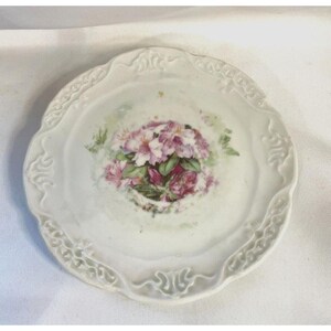 May include: A white porcelain plate with a decorative floral design. The plate features a cluster of pink flowers with green leaves, set against a white background. The plate has a scalloped edge with a raised design.