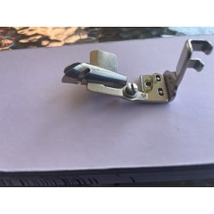 May include: A silver sewing machine foot with a curved needle guide and a screw for attachment.