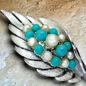 May include: A silver-toned metal brooch shaped like a feather or leaf, featuring a cluster of round, smooth turquoise-colored beads and white pearls set along the center spine of the textured metallic structure.